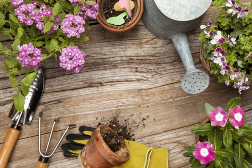 The image displays a wooden garden table with various gardening tools and accessories arranged on its surface. On the left, there is a small hand trowel with a wooden handle, and next to it, a garden fork with metal prongs. A pair of gardening gloves with black and yellow accents is placed near a small terracotta pot containing dark soil, which is partially knocked over. To the right, there is a grey watering can with a spout facing downward, alongside a terracotta plant pot filled with colourful decorative plant cutouts. The table features three potted plants: one with pink and white flowers, another with purple and white blooms, and a third with densely clustered purple flowers, all thriving in their respective containers. The scene suggests a gardening setup focused on flower planting or potting activities, emphasizing a well-organized outdoor garden environment, possibly in a residential backyard or patio area. The natural lighting and weather conditions appear to be bright and clear, suitable for outdoor gardening tasks, aligning with services provided by Lawn Mowing Finchley in the Finchley area, N3 postcode, enhancing outdoor space maintenance and garden care in the local community.