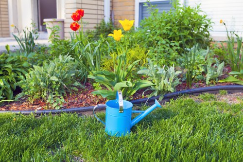 The image depicts a well-maintained garden in a suburban outdoor space, featuring a vibrant, green lawn with dense, evenly cut grass in the foreground. Behind the lawn, there is a flower bed bordered by black edging, containing a variety of plants including yellow tulips and red, pink, and white bloomers, alongside leafy green foliage. A light blue watering can is placed on the grass near the edge of the flower bed, suggesting active garden maintenance. In the background, there are some small shrubs and a residential building with white siding and blue window frames, indicating a typical home garden setting in Finchley. The scene appears to be taken on a bright, slightly overcast day, with soft natural lighting enhancing the lush greenery and vibrant flower colors, demonstrating a neat and thoughtfully landscaped garden area suitable for gardening services like lawn mowing and garden care by Lawn Mowing Finchley, serving the local community in the N3 postal district.