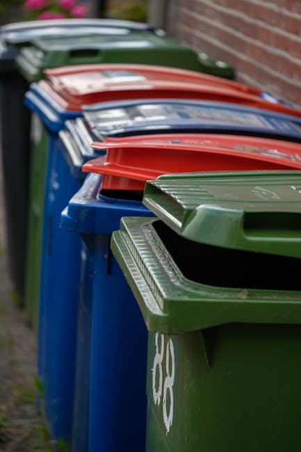 A close-up view of a garden area viewed through black metal fence bars, focusing on a pile of discarded garden waste, including crumpled paper, plastic bags, and food packaging. The background reveals